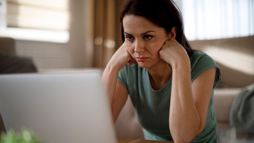 girl struggling with hands under chin, seems stressed out and has computer in front of her. Green top, dark haired