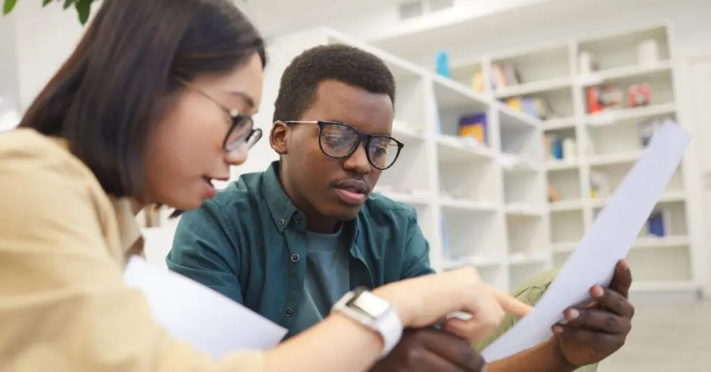 African American Student, Holding Piece of White Paper, Sitting Next to an Asian female who is pointing at the paper seeming to instruct a focus of one specific portion.